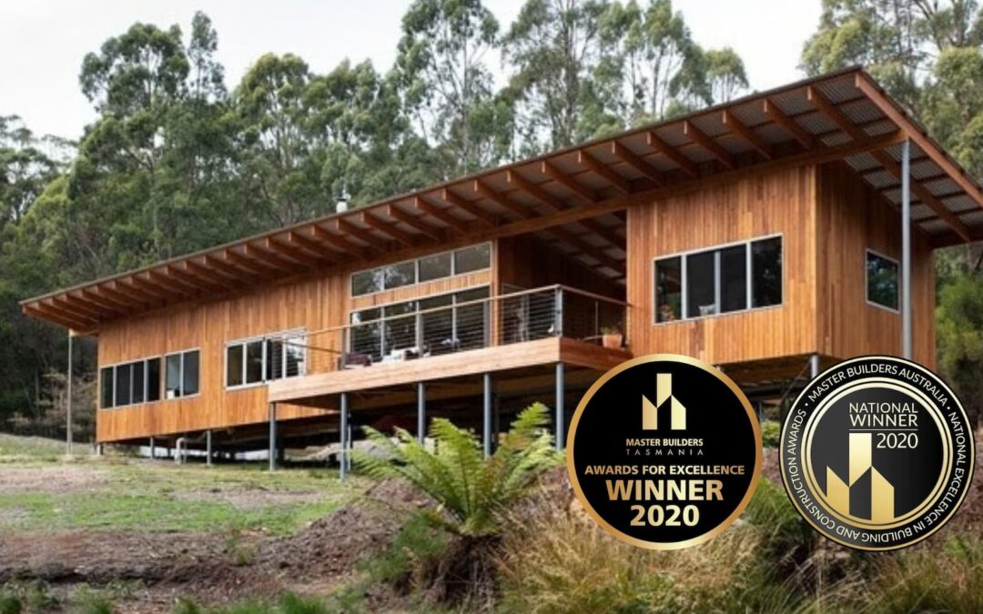 Modern wooden house with large windows and a deck surrounded by trees. Two circular badges display “Master Builders awards 2020.”.