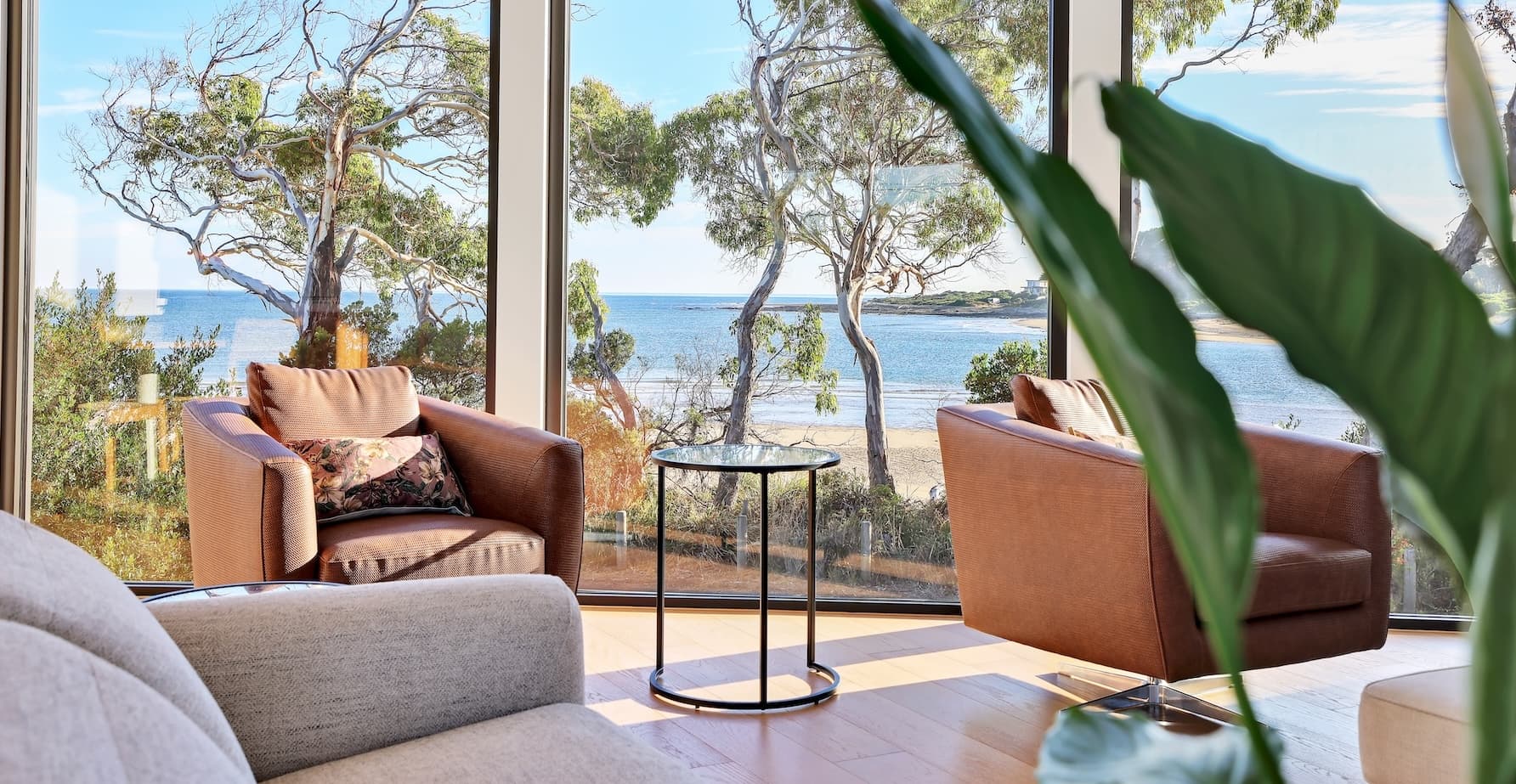 A modern living room features two brown armchairs facing expansive windows, offering stunning beach and tree views. Between them sits a sleek round glass coffee table, while a lush plant with large green leaves adds a touch of nature.