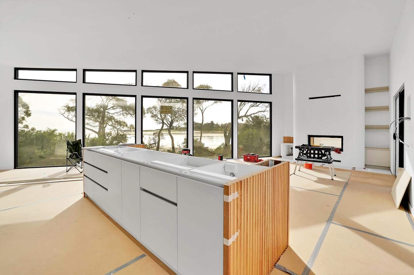 Modern living area under construction. An unfinished kitchen island is in the middle of the room, complemented by a fireplace to the right and minimal furnishings. Walls and floor remain partly completed.