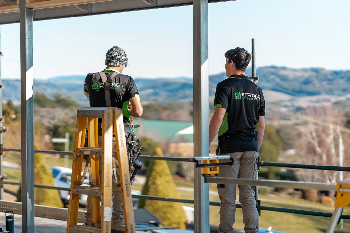 Adam and his employee in black shirts with Strong Constructions embroidered on the back stand on a building. One is behind a ladder. There is a scenic landscape with hills in the background. Various tools are visible around the site.