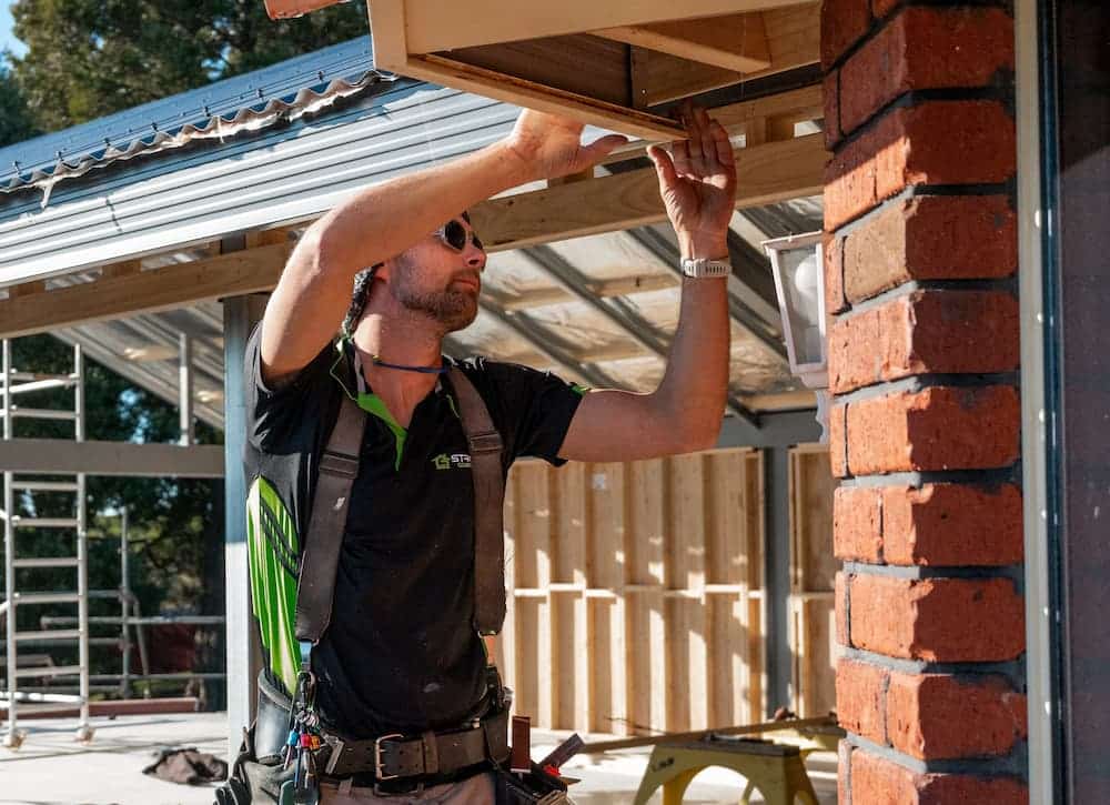 Adam Armstrong wearing a black shirt, with the logo, Strong Constructions embroidered on the right hand side. Adam is wearing sunglasses, and a tool belt. Adam i s working under s roof section with a brick wall and an unfinished wooden frame in the background.
