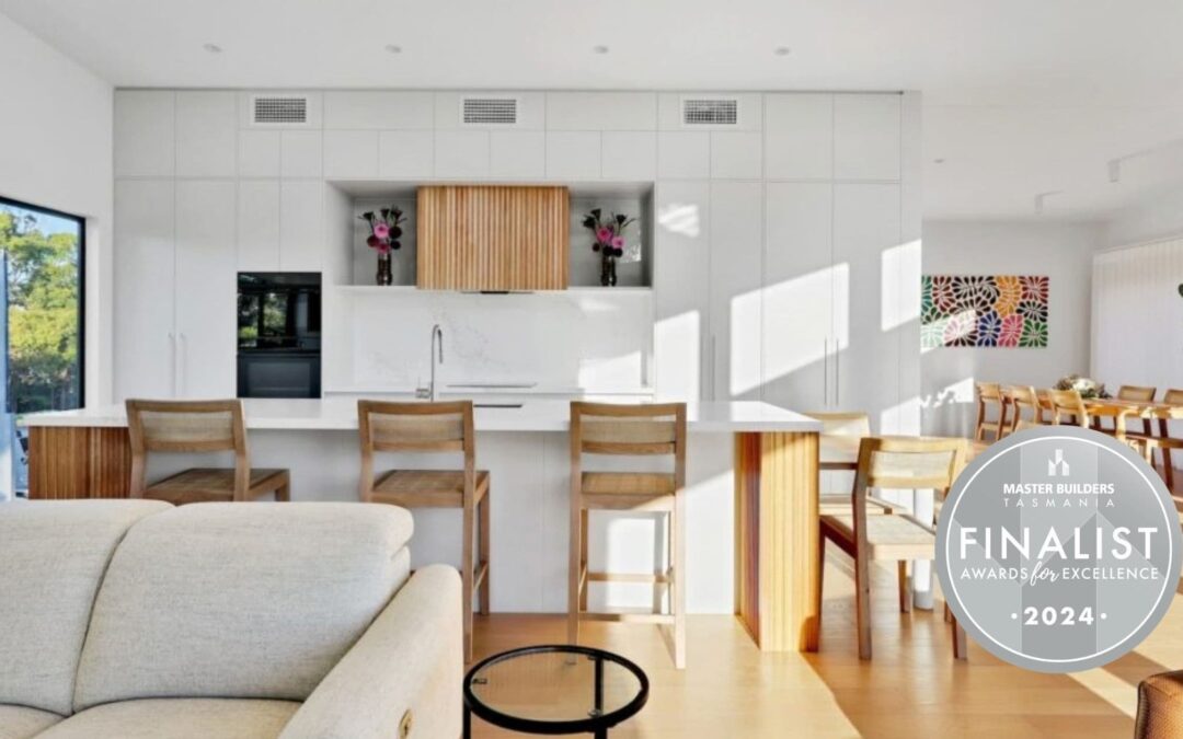 Modern kitchen with white cabinetry, light wood accents, an island with barstools, and a 2024 Master Builders Tasmania Finalist award badge in the lower right corner.
