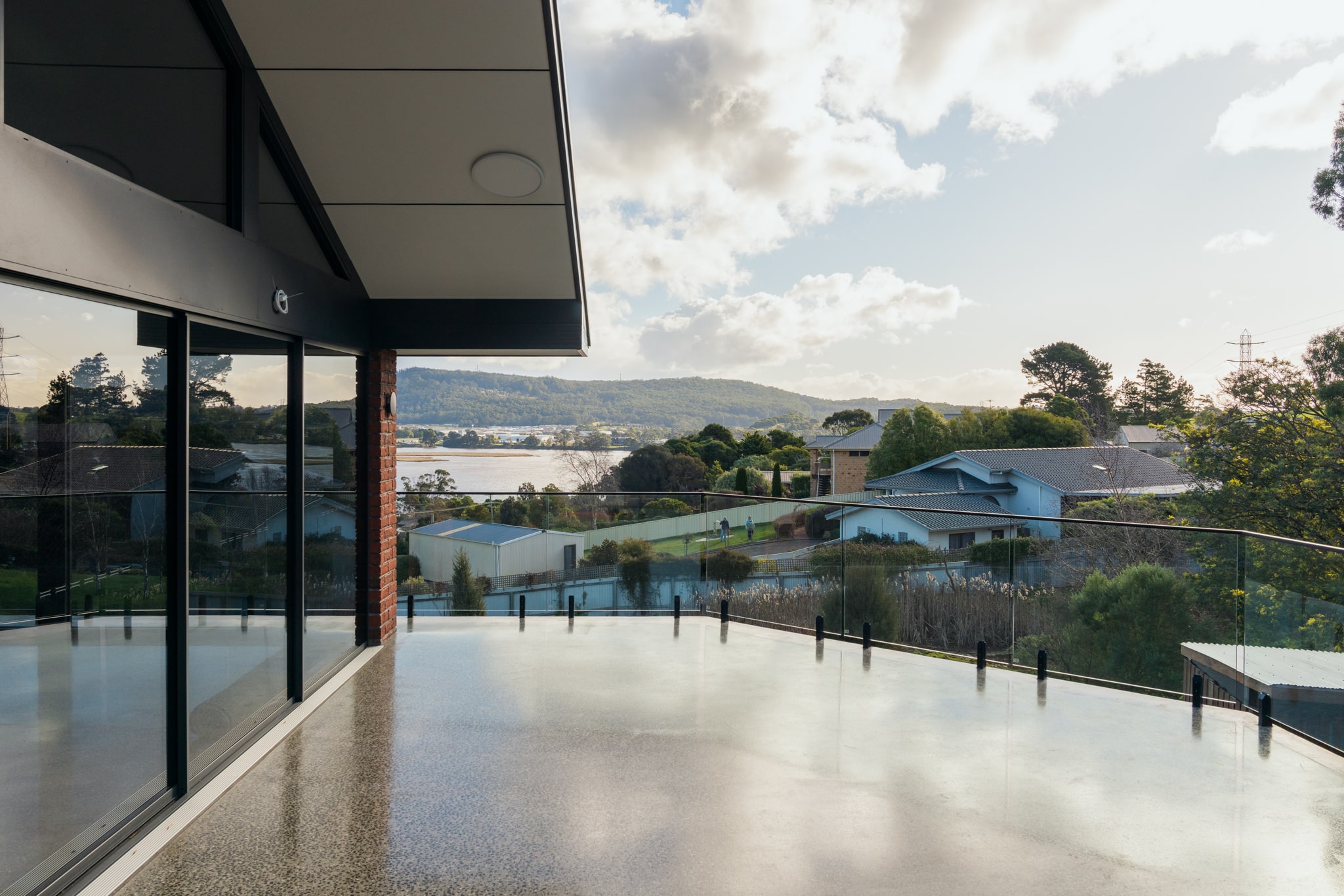 Copy of 21 Highfield Rd (1) Modern house balcony with glass railing overlooks residential neighborhood, trees, and a lake with hills in the background under a partly cloudy sky.