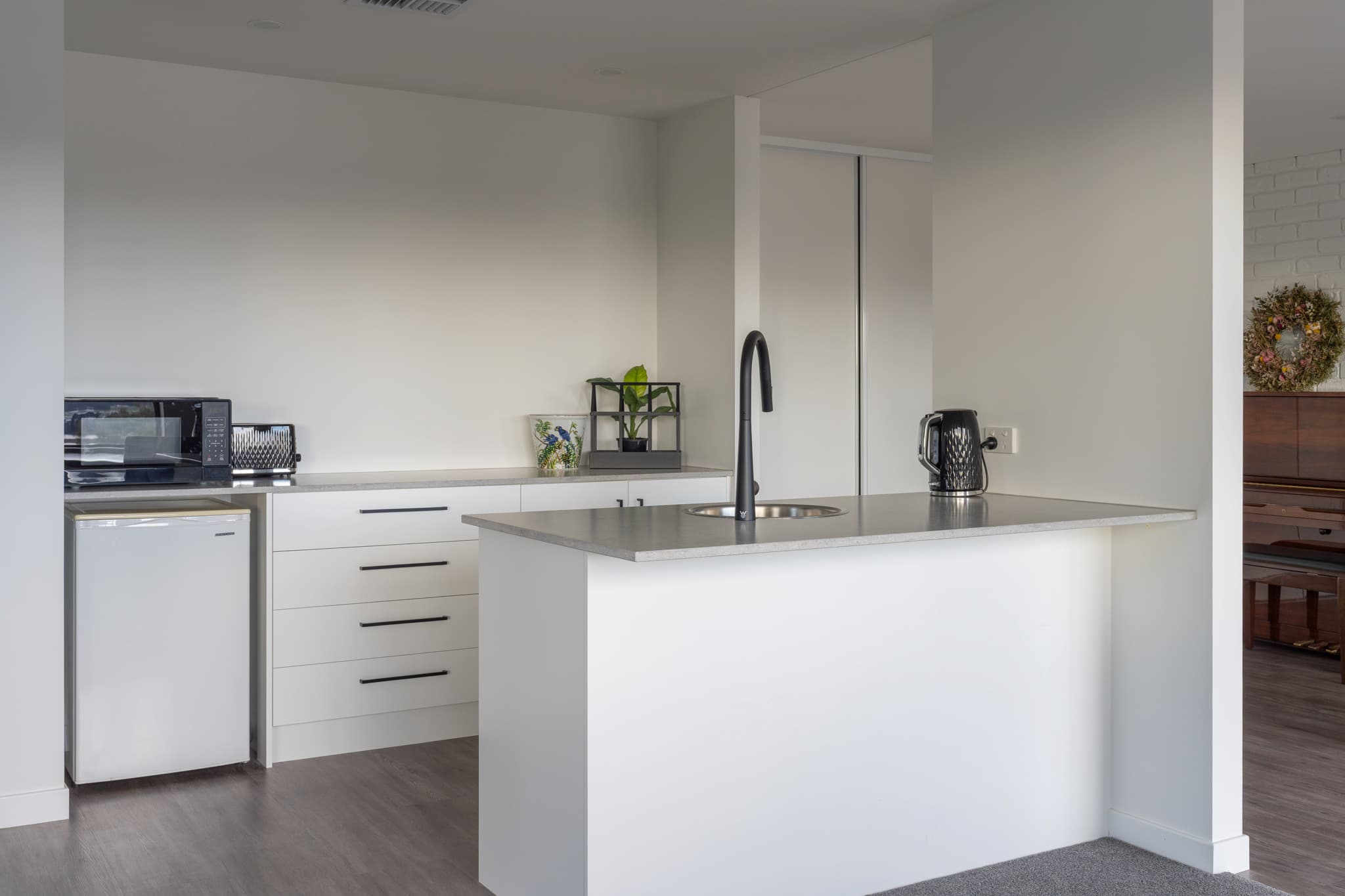 Modern kitchen with white cabinets, a small fridge, microwave, toaster, black faucet, and a kettle on the countertop; piano visible in adjacent room.
