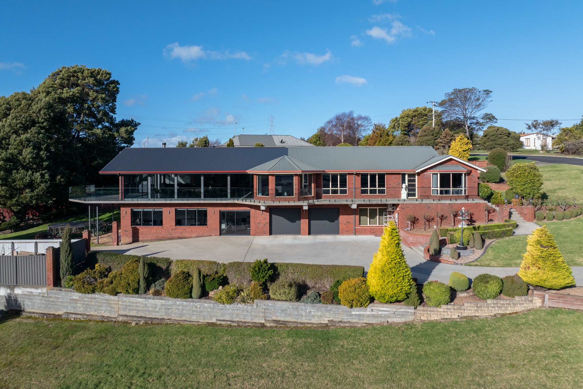 Large two-story brick house with a dark roof, multiple windows, and a spacious driveway, surrounded by neatly landscaped lawns and shrubs under a clear blue sky.