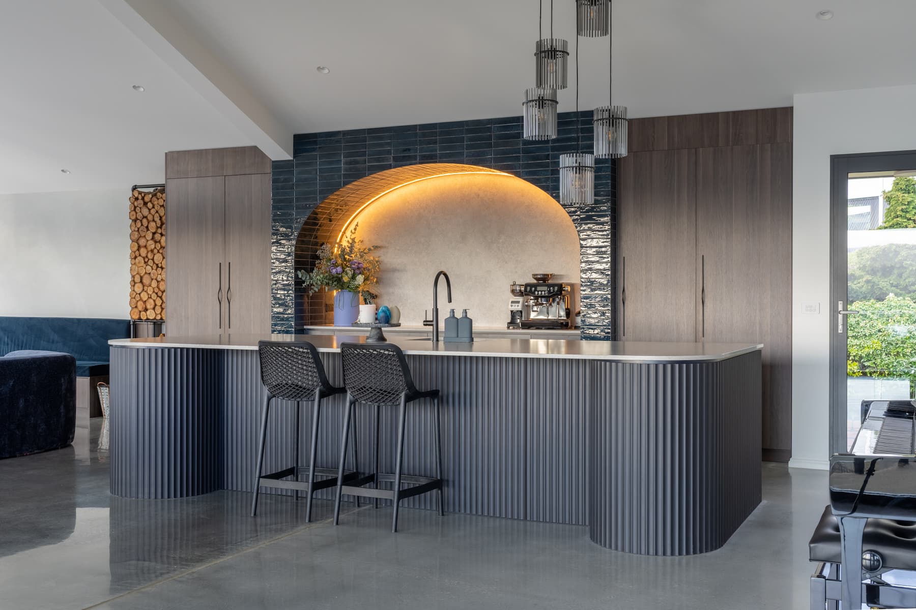 Copy of 21 Highfield Rd-15 (1) (1) Modern kitchen with dark cabinetry, a curved island, two black bar stools, arched backsplash with accent lighting, and built-in appliances on a polished concrete floor.