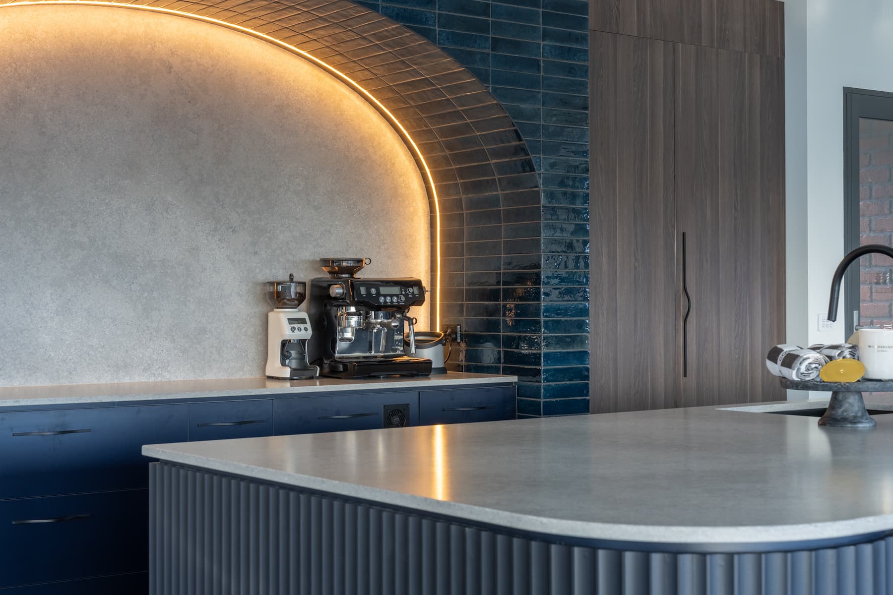 Modern kitchen with a concrete countertop, espresso machine, dark blue cabinets, blue tile backsplash, wood cabinetry, and arched LED lighting feature.