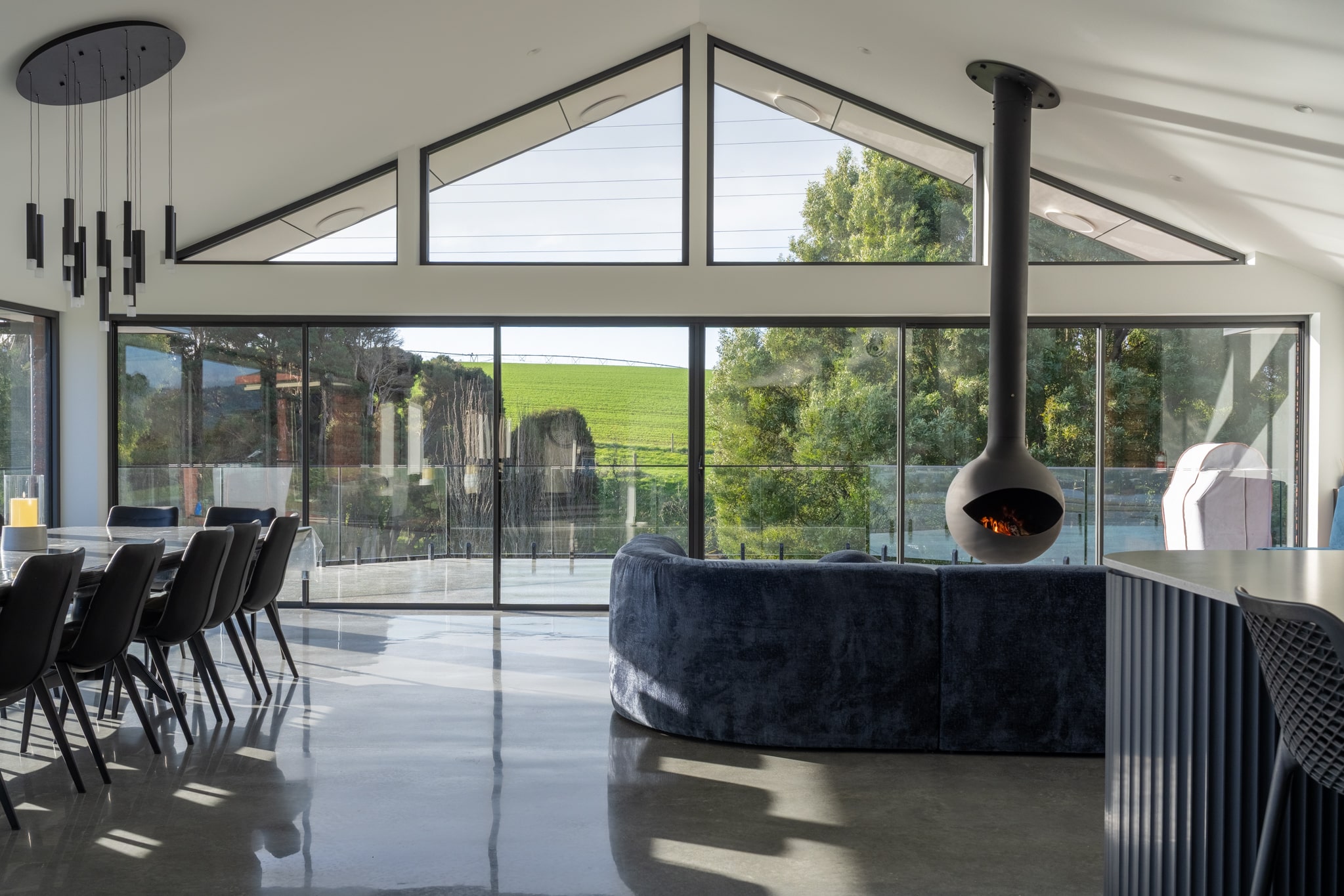 Modern open-plan living and dining area with large glass windows, a suspended fireplace, blue sofa, and views of green landscape outside.