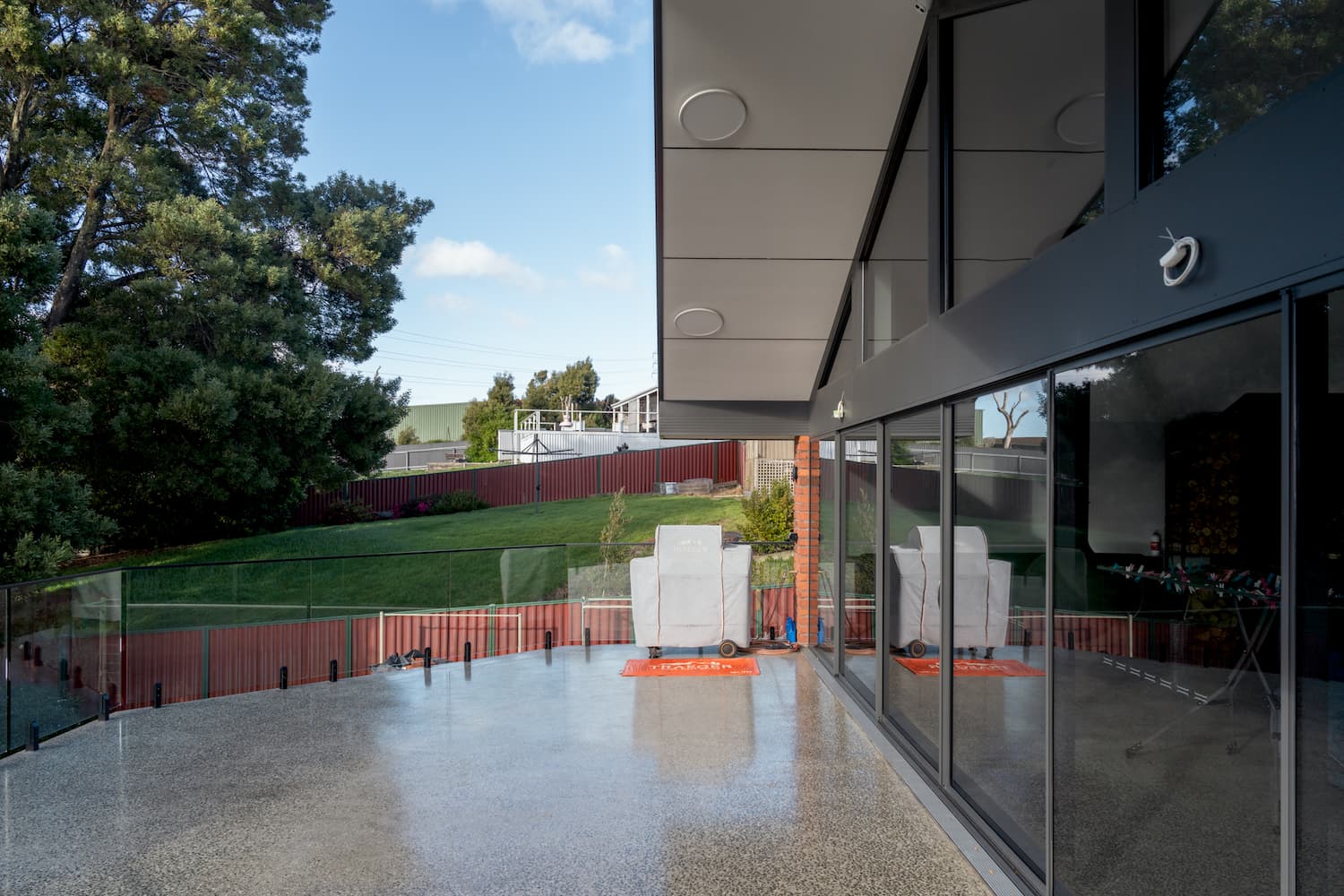 Modern outdoor patio with glass fencing, covered grill, two covered chairs, and a view of a grassy yard and trees, seen through large glass windows.