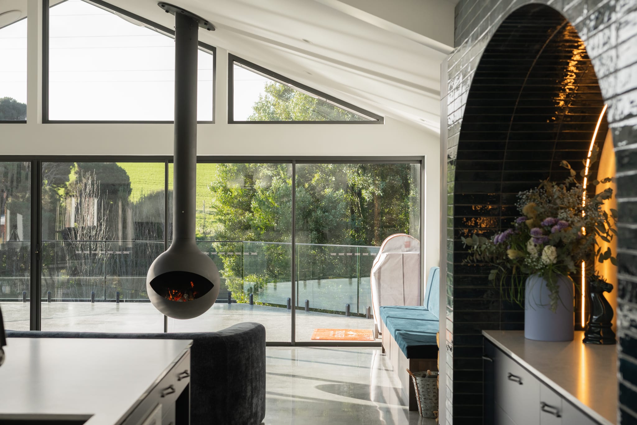 Modern living space with a suspended black fireplace, large windows, outdoor view, built-in bench seating, and a kitchen area with a flower arrangement on the counter.