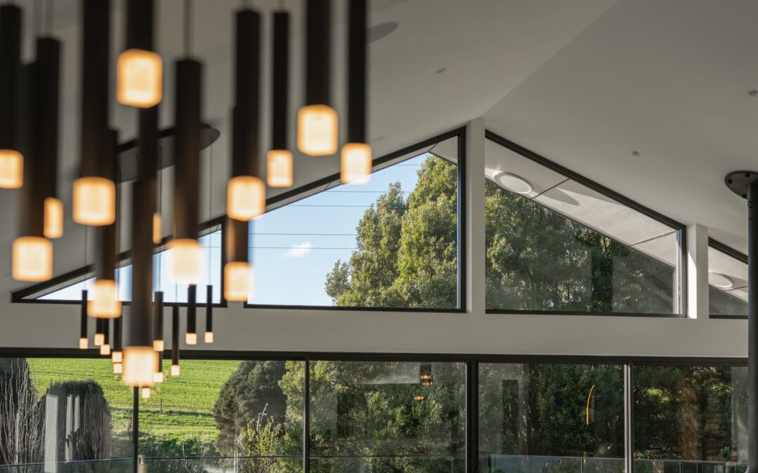 Modern interior with hanging pendant lights, large glass windows, and a view of green trees and sky outside. Geometric ceiling design with natural light entering the space.
