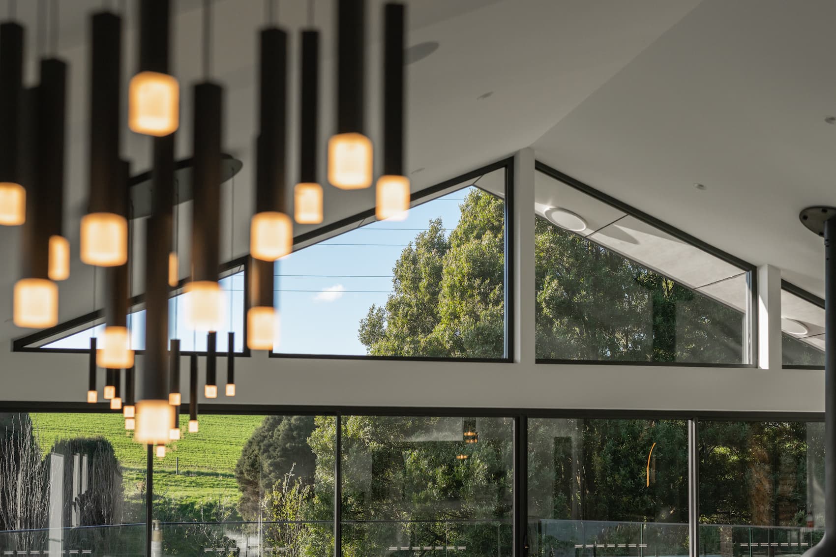 Modern interior with hanging pendant lights, large glass windows, and a view of green trees and sky outside. Geometric ceiling design with natural light entering the space.