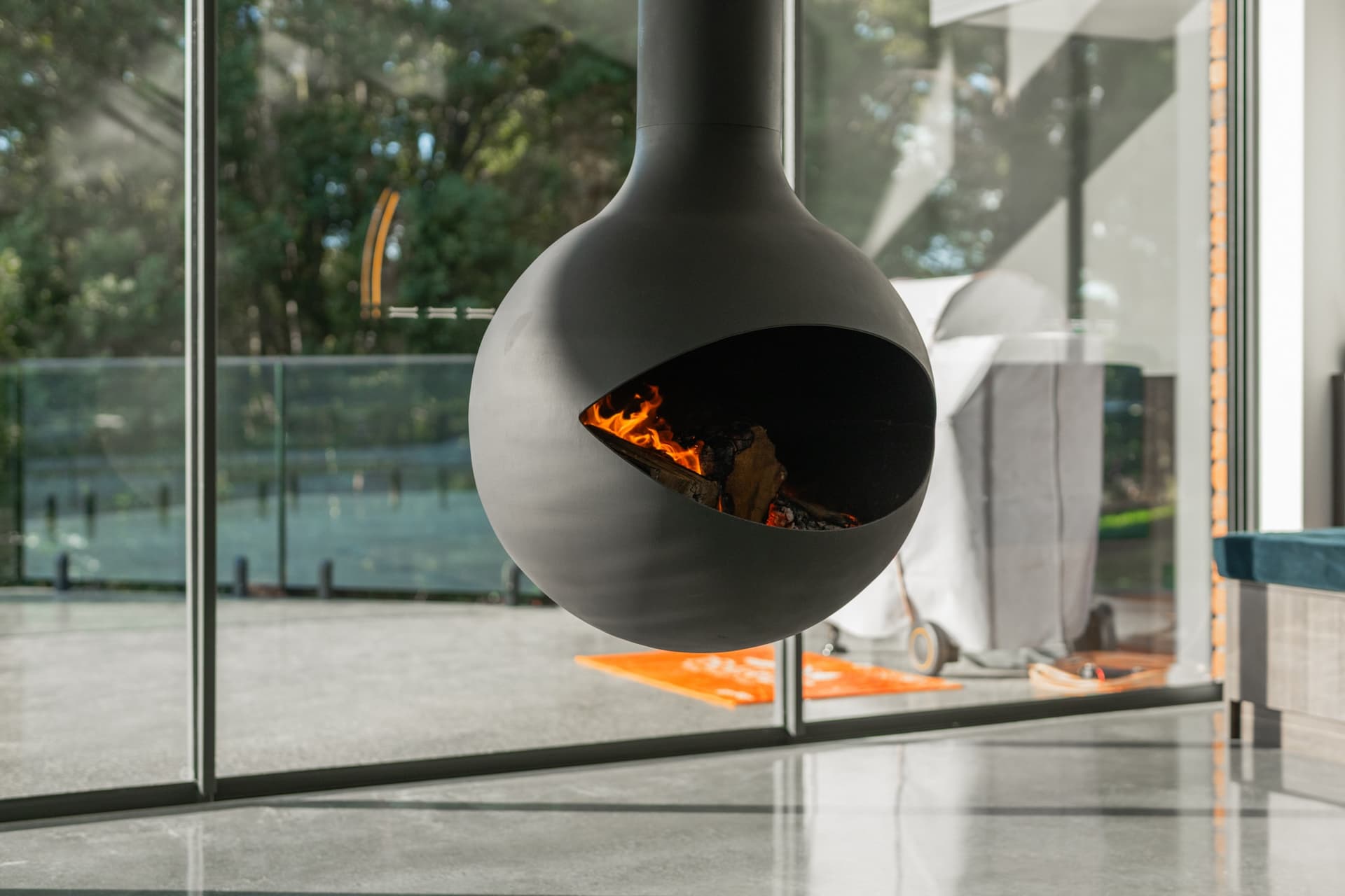 A modern black suspended fireplace with a fire burning inside, positioned indoors near large glass windows overlooking a patio and trees outside.