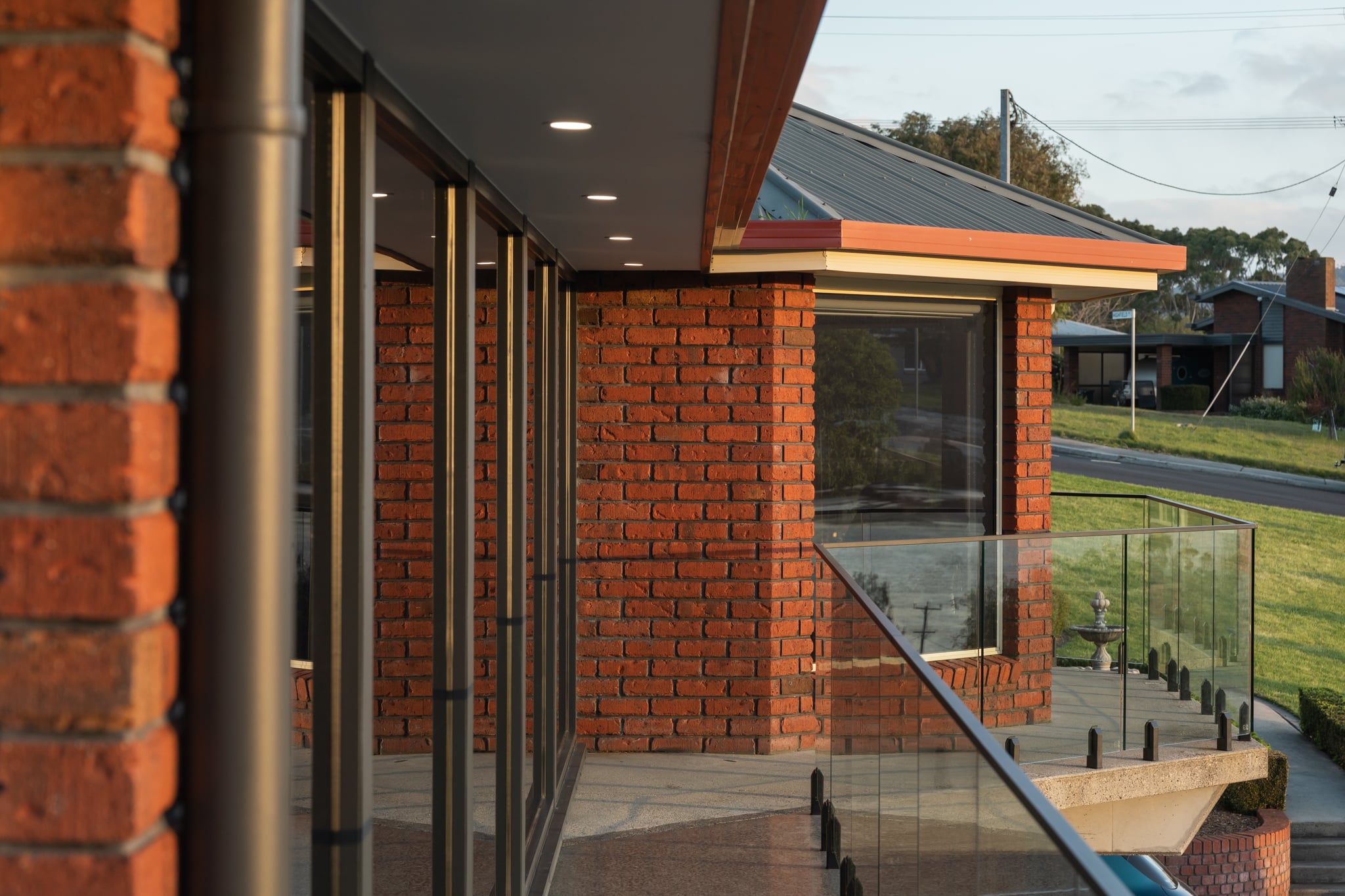A modern building with red brick walls, large glass windows, and a glass balcony railing, overlooking a grassy area and a residential street.