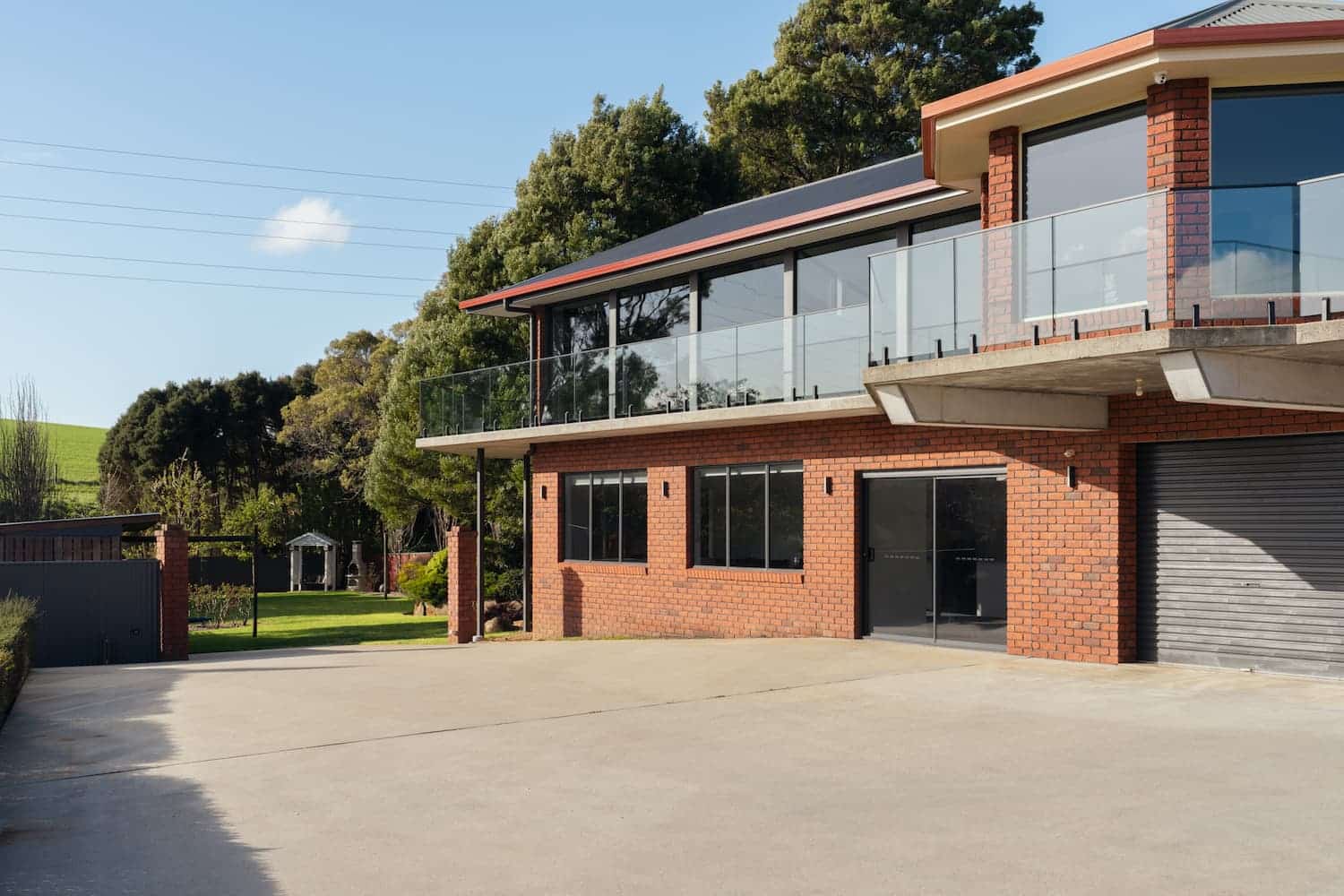 A modern two-story brick house with large glass windows, a balcony, and a spacious concrete driveway surrounded by greenery and trees.