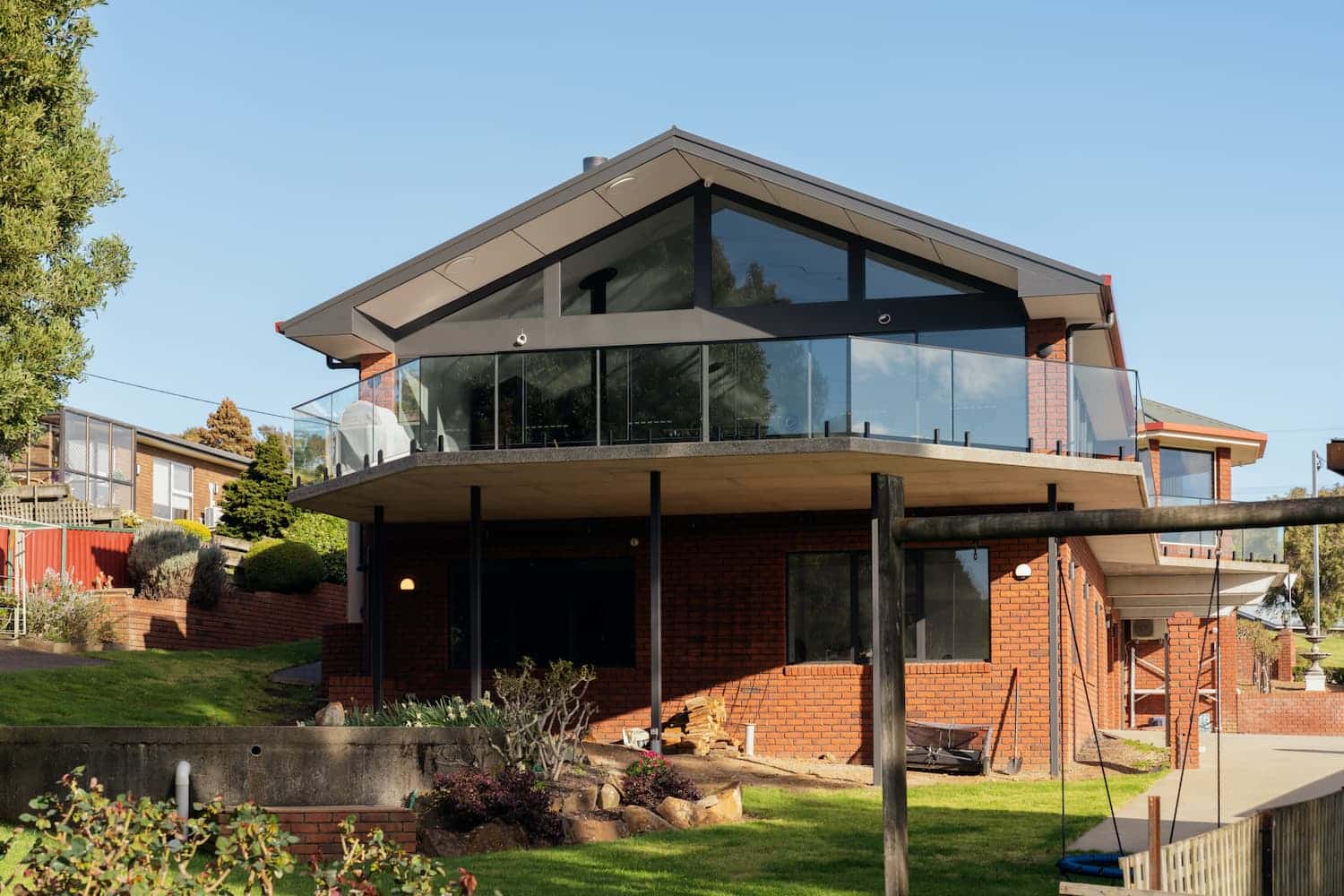 A modern two-story brick house with large glass windows and a wraparound balcony, set on a landscaped lawn under a clear sky.