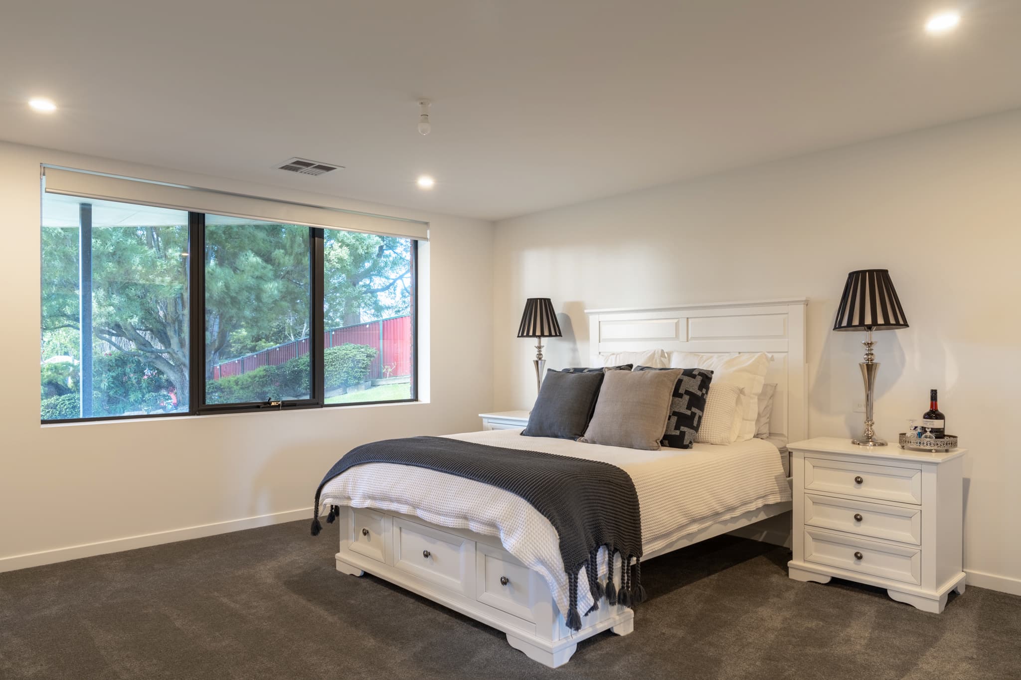A modern bedroom with a large bed, white furniture, two bedside tables with lamps, and a window overlooking greenery. The room has neutral tones and minimal decor.