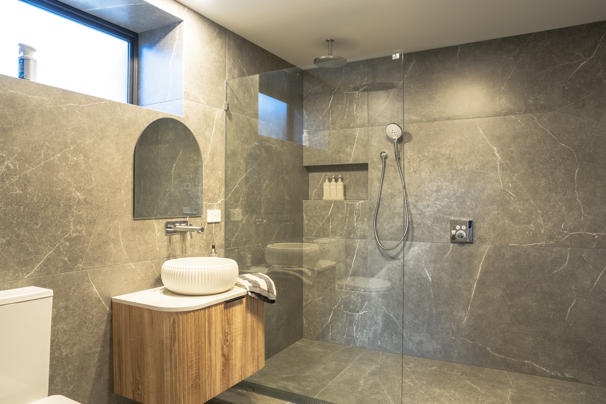 Modern bathroom with gray stone tiles, a floating wooden vanity with a round white sink, a frameless glass shower enclosure, and a wall-mounted rainfall showerhead.