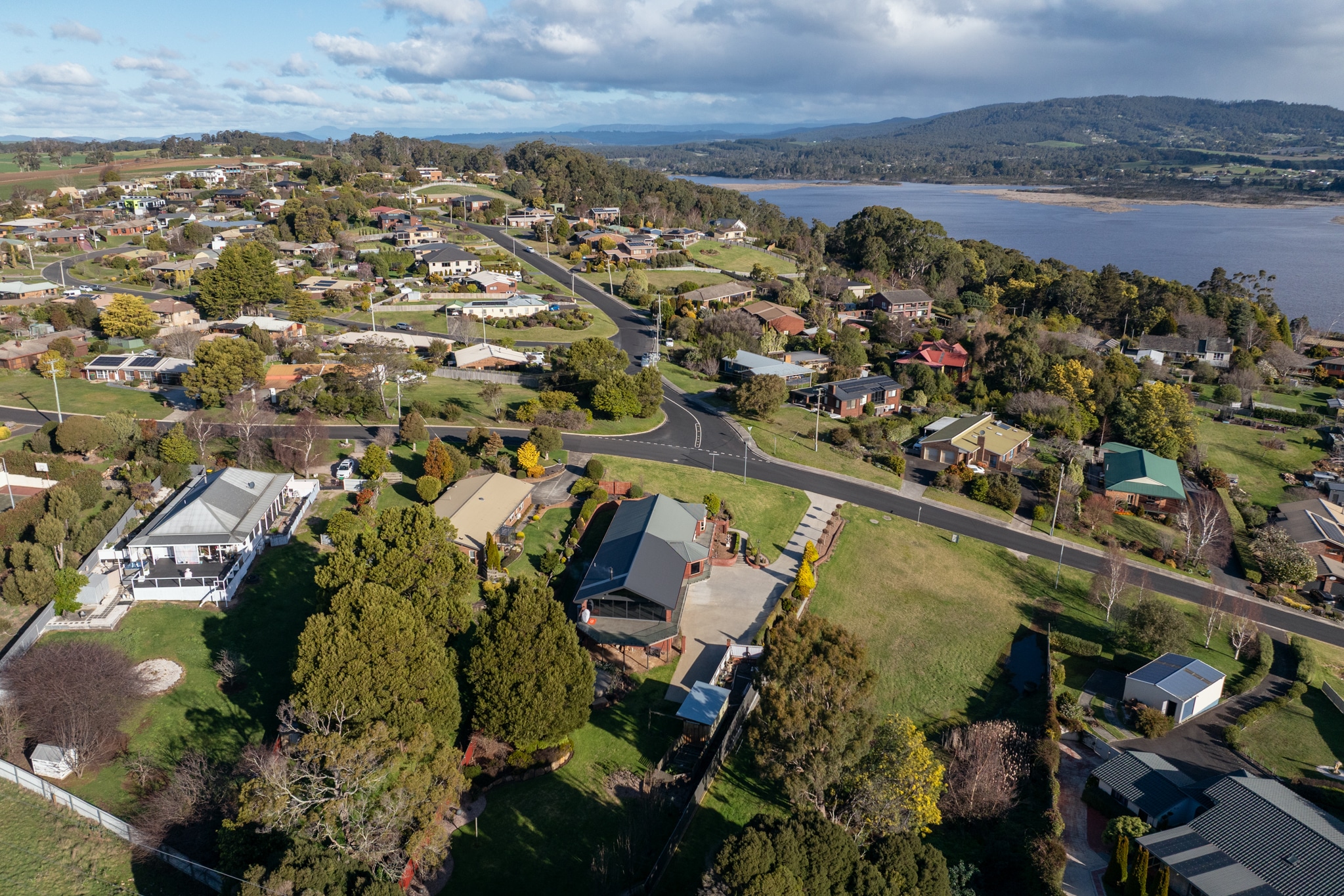 Aerial view of a suburban neighborhood with houses, trees, winding roads, and a large body of water in the background under a partly cloudy sky.