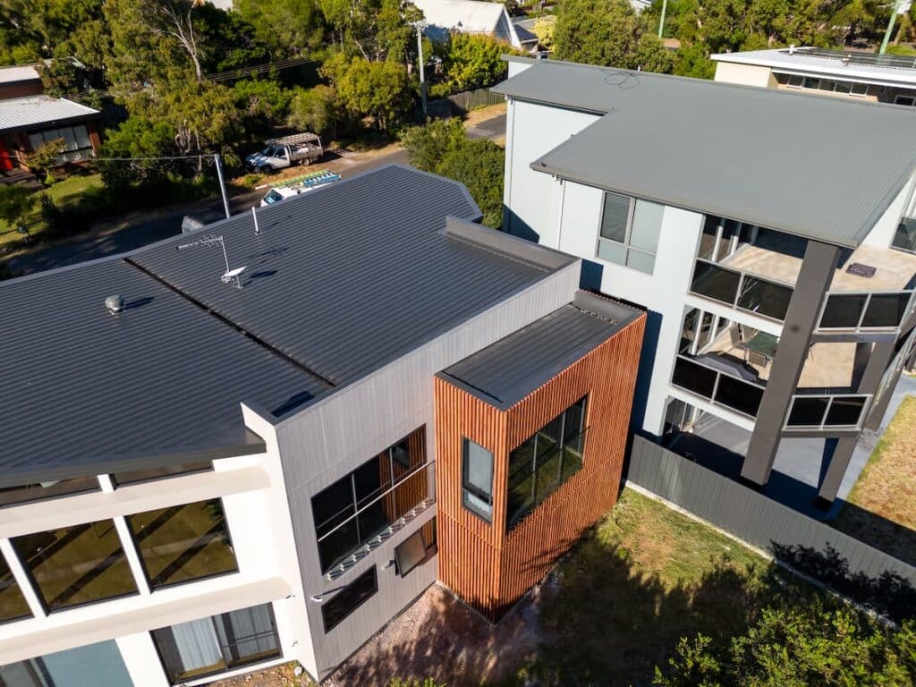 Aerial view of two modern multi-story houses with flat roofs and large windows, separated by a fence and surrounded by trees.