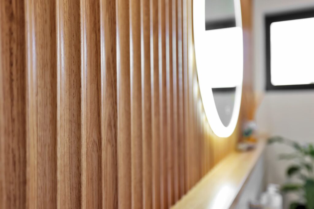 Close-up of vertical wooden slats on a wall with part of a round, illuminated mirror and a blurred window in the background.