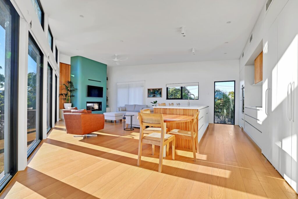 Modern open-concept living and dining area with large windows, light wood floors, minimalist furniture, and a green accent wall with a mounted TV.