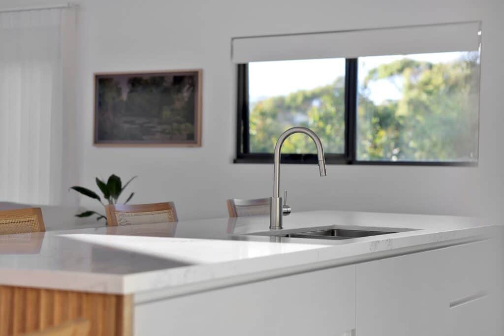 Modern kitchen with a white marble countertop, a stainless steel faucet, wooden chairs, a plant, and a window letting in natural light.