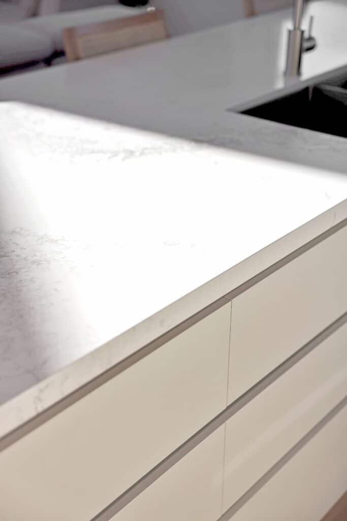 Close-up of a white marble kitchen countertop with subtle grey veining, next to a sink, with sunlight casting shadows across the surface.