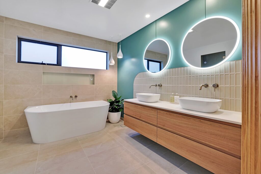 Modern bathroom with a freestanding bathtub, double vanity with vessel sinks, two round backlit mirrors, and beige and teal wall tiles. A potted plant sits beside the tub.