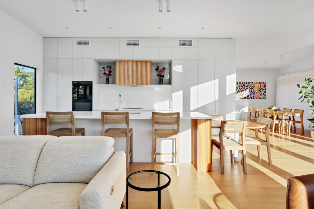 Modern open-plan kitchen and dining area with white cabinetry, wooden stools, a large island, and bright natural light streaming in through windows.