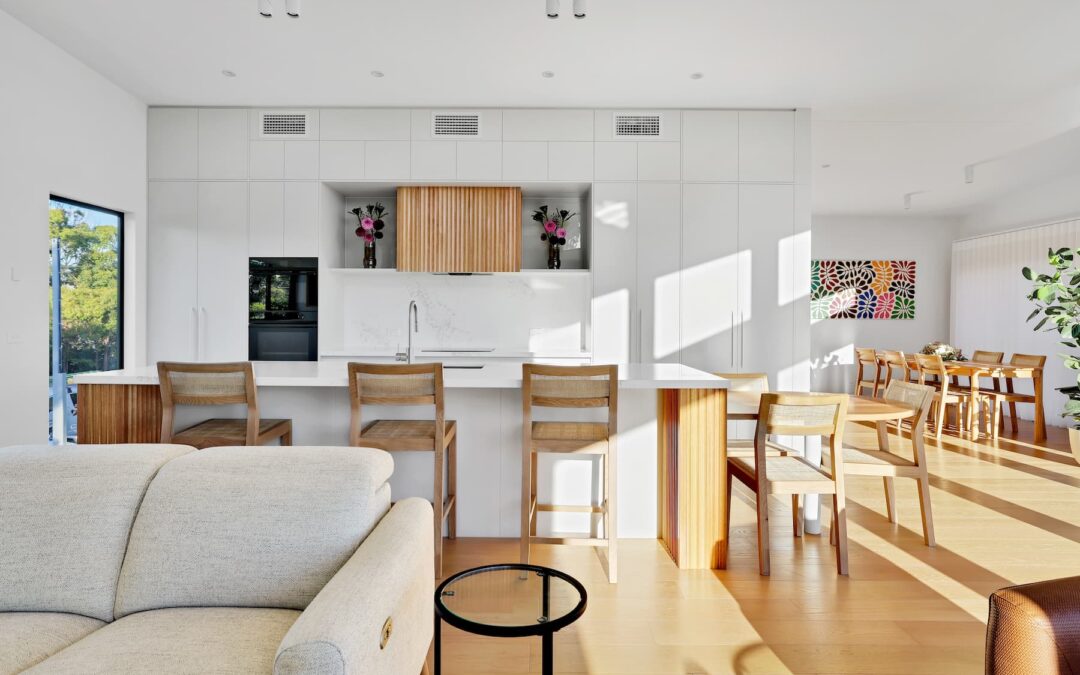 Modern open-plan kitchen and dining area with white cabinetry, wooden stools, a large island, and bright natural light streaming in through windows.