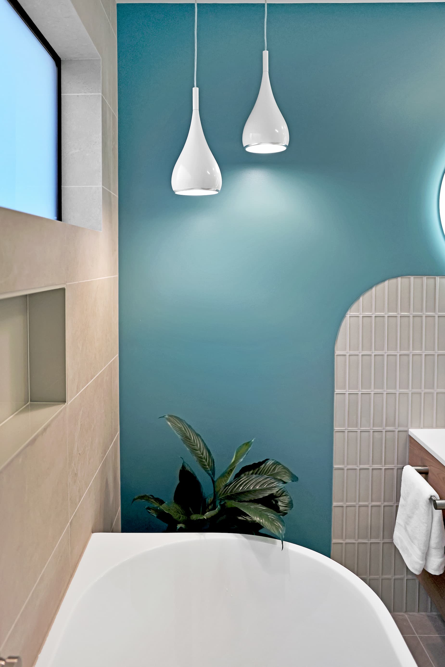 SC Turners Beach i-31 (1) Modern bathroom with a blue accent wall, two white pendant lights, a plant next to a white bathtub, and beige tiled surfaces.