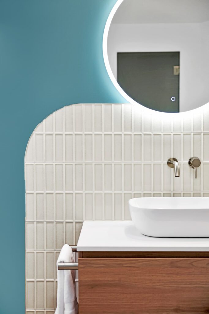 Modern bathroom with a round illuminated mirror, white rectangular sink, wall-mounted faucet, wood vanity, and blue and cream tiled wall.