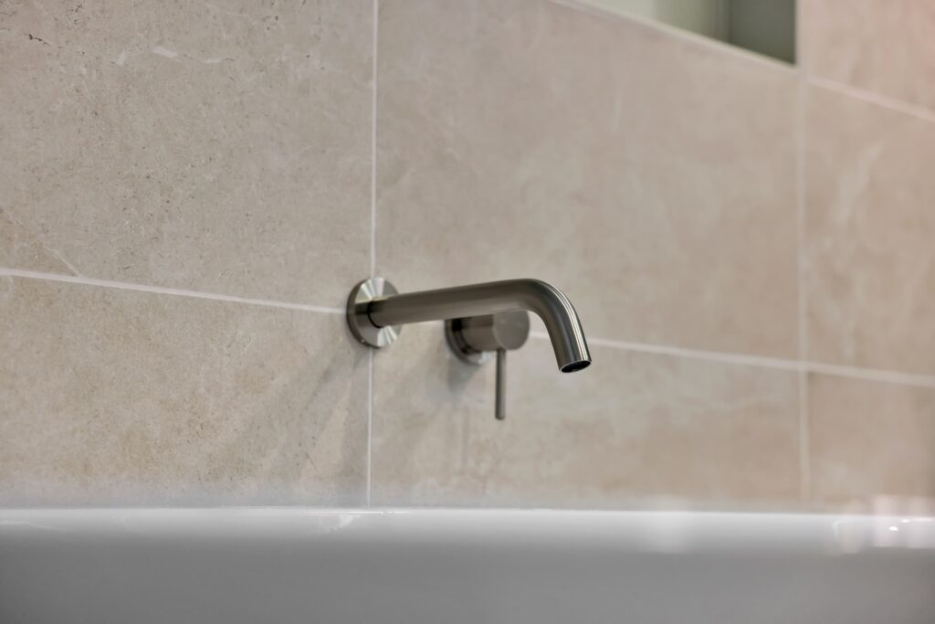 A stainless steel wall-mounted faucet with a single lever handle installed above a white sink against beige tiled walls.