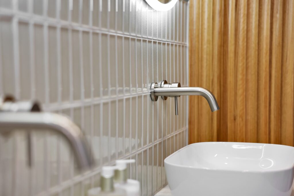 Close-up of a modern bathroom with a wall-mounted faucet above a white square sink, beige tiled wall, and blurred toiletry bottles in the foreground.