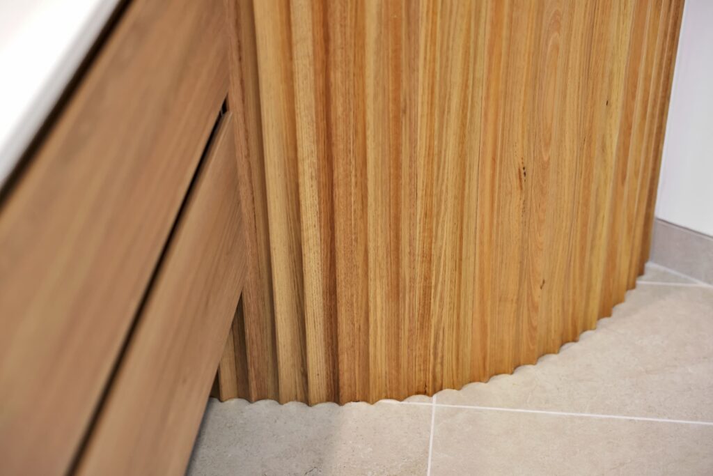 Close-up of a wooden cabinet with vertical grooves and scalloped edge at the bottom, next to a tiled floor and another wooden drawer unit.