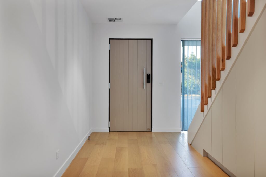 Modern entryway with light wood flooring, a beige front door, white walls, and a staircase with wooden handrails on the right. Natural light comes in from a window near the stairs.