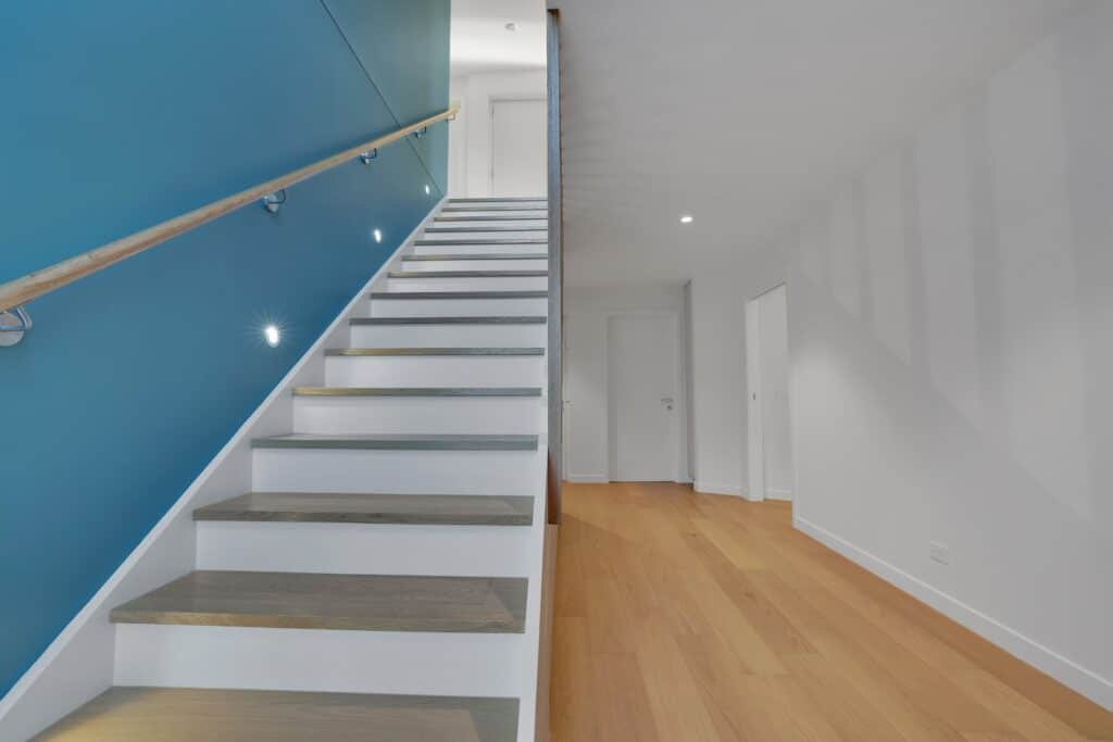 Modern interior with a wooden staircase featuring a blue accent wall and built-in lights, next to a hallway with light wood flooring and white walls.