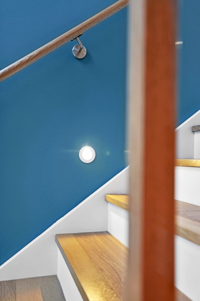 Wooden stairs with white risers beside a blue wall, featuring a round wall light and a metal handrail.