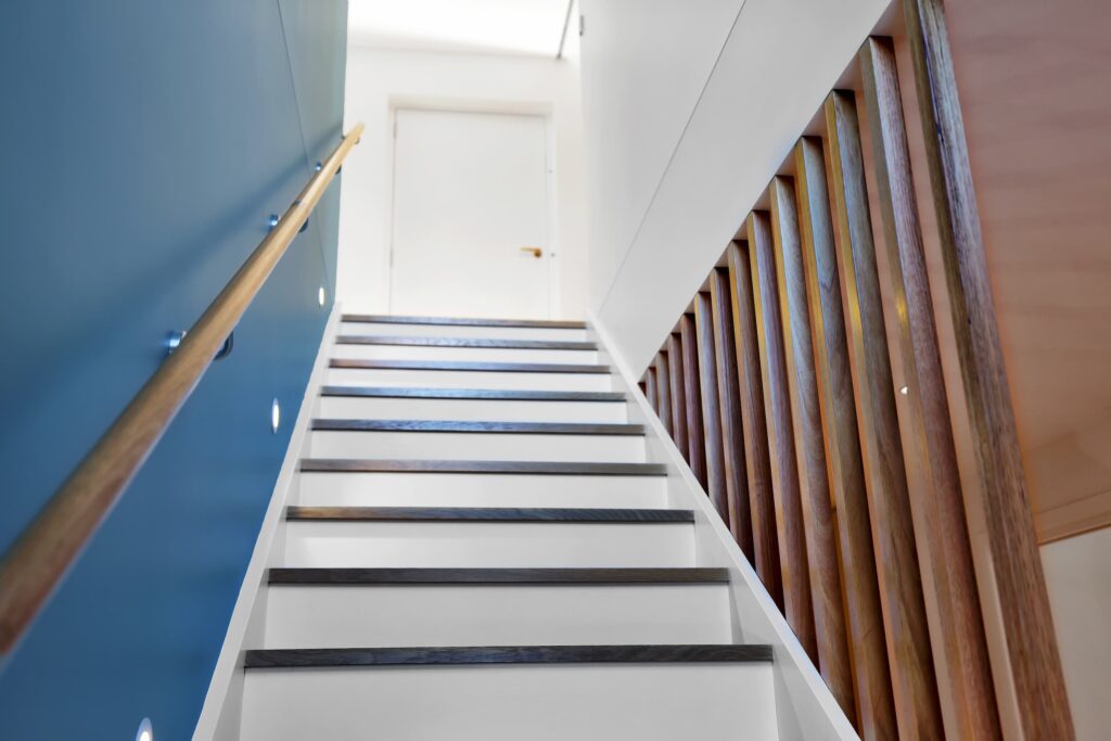 View of a modern indoor staircase with white risers, dark treads, a blue wall with handrail and lights on the left, wooden vertical balusters on the right, and a closed door at the top.
