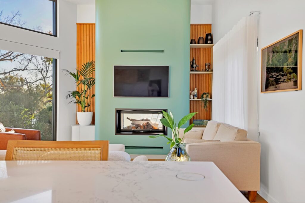 Modern living room with a green accent wall, built-in fireplace, wall-mounted TV, beige sofa, large windows, and wood paneling with decorative shelves.