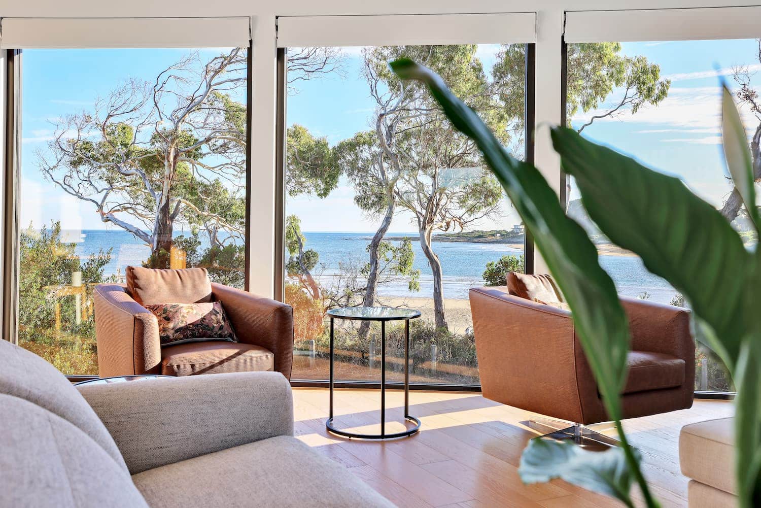 SC Turners Beach i-6 (1) A modern living room with large windows overlooks a beach and trees. Two brown armchairs and a small round table face the view; a plant is visible in the foreground.