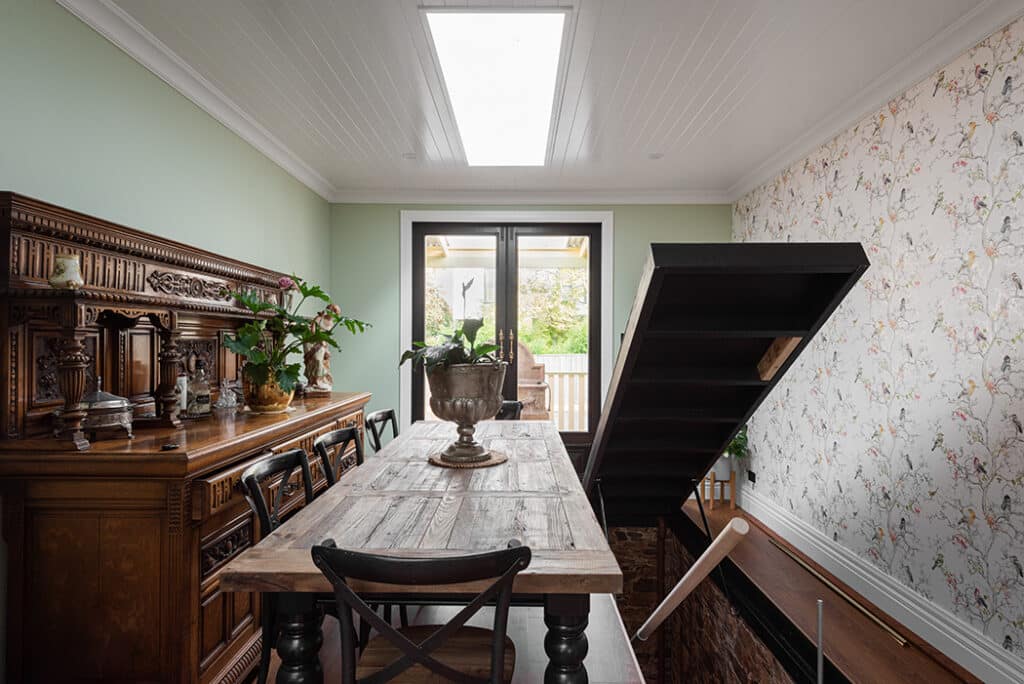 A dining room with a wooden table and chairs, an ornate sideboard, floral wallpaper, and an open staircase leading down, with a glass door and skylight providing natural light.