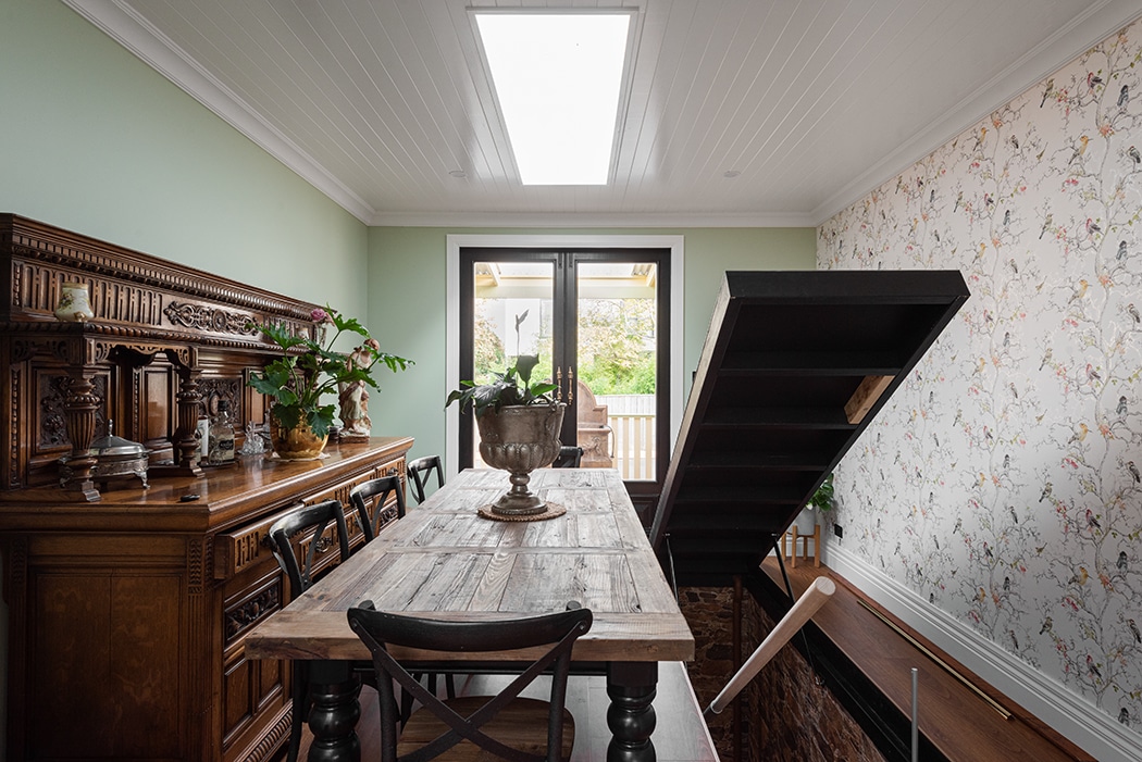 A dining room with a wooden table and chairs, an ornate sideboard, floral wallpaper, and an open staircase leading down, with a glass door and skylight providing natural light.