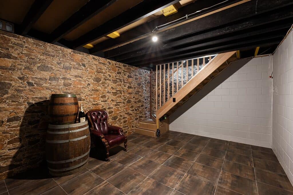 A basement with tiled floor, exposed ceiling beams, a stone and cinder block wall, a wooden staircase, a leather chair, and two stacked barrels.