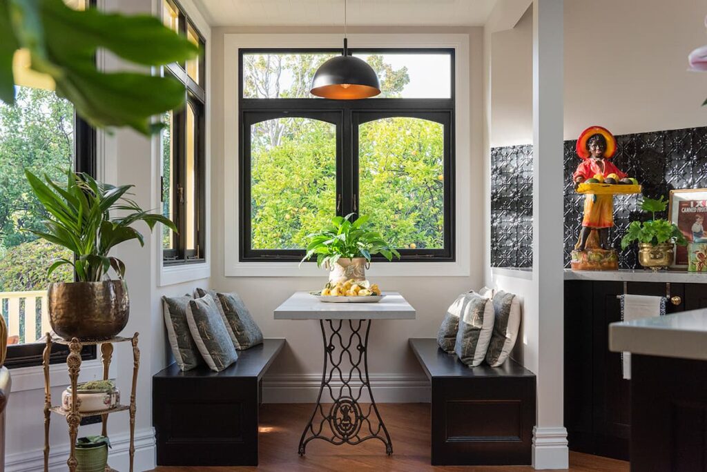 A small dining nook with a square table, two bench seats with cushions, potted plants, and a large window letting in natural light.