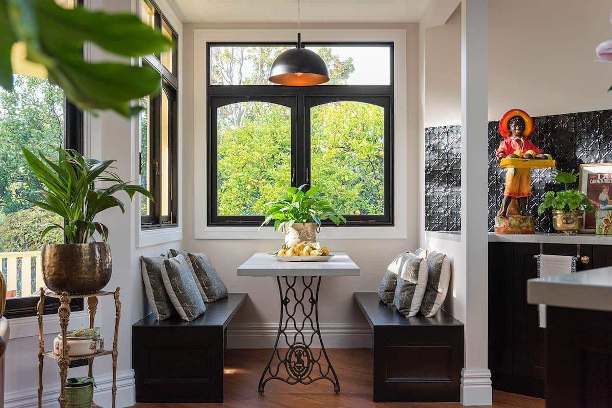 A small dining nook with a square table, two bench seats with cushions, potted plants, and a large window letting in natural light.