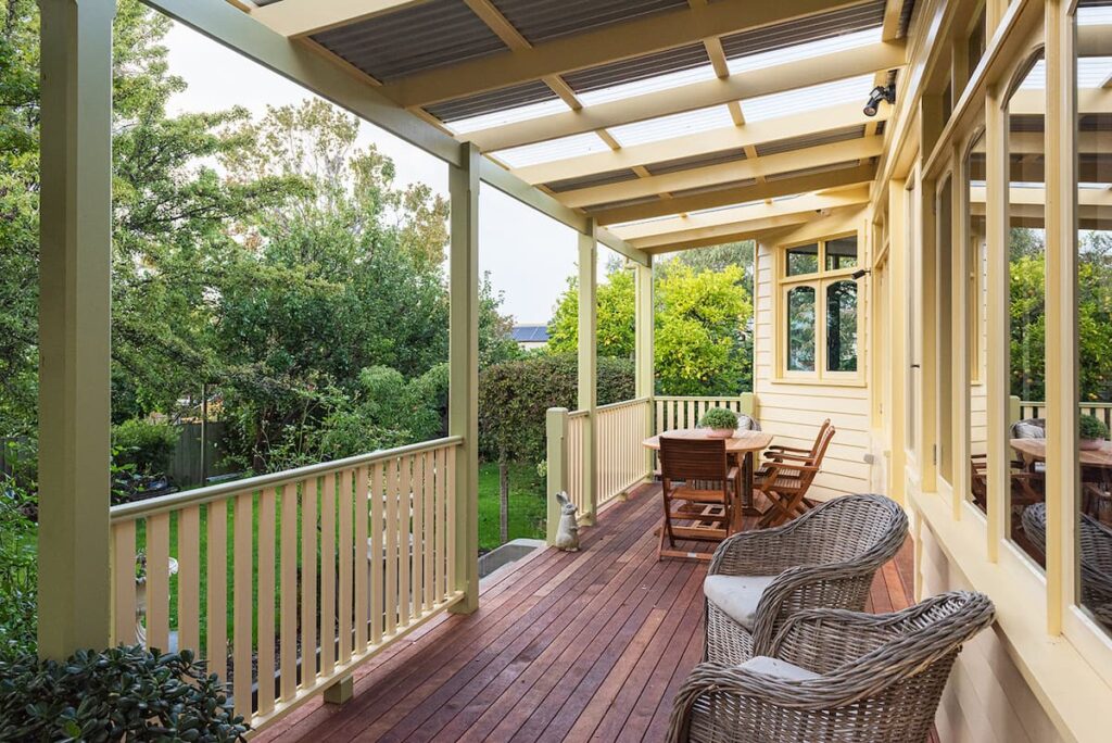 Covered wooden porch with wicker chairs, a dining table, and chairs, overlooking a green backyard with trees and shrubs on a sunny day.
