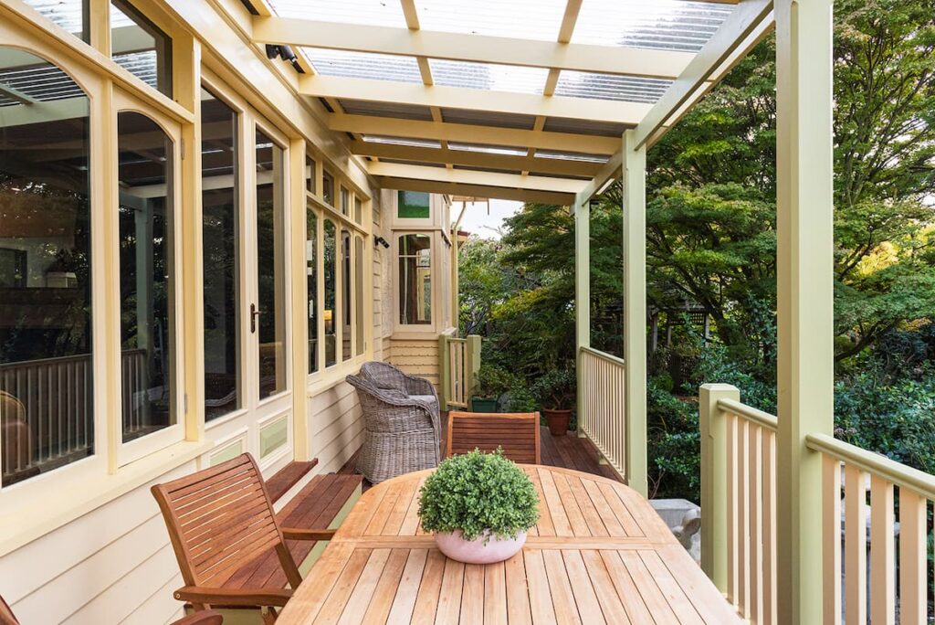 Wooden patio with a clear roof, outdoor table and chairs, a potted plant centerpiece, and garden foliage visible in the background.
