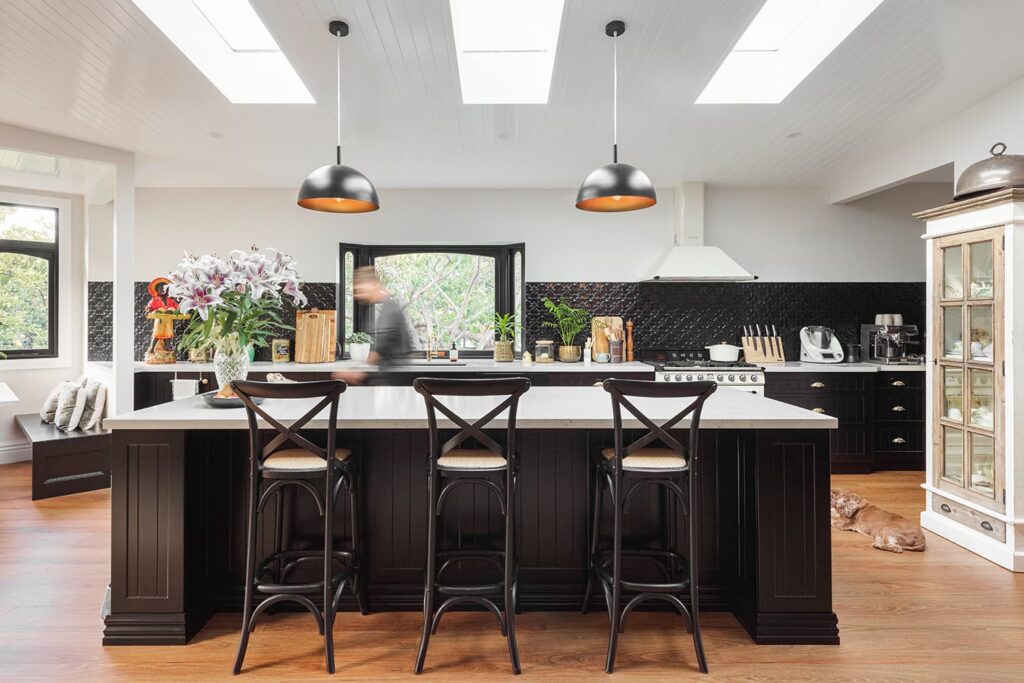 Modern kitchen with black island, three barstools, hanging lights, plants, and kitchen appliances; a person is blurred in motion and a dog lies on the wooden floor.